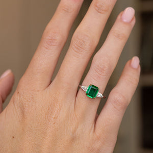 Emerald-cut three-stone engagement ring worn on the hand, showing the vivid green gemstone and elegant trapezoid diamond shoulders.