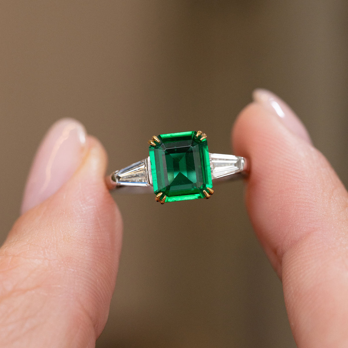 Handheld close-up of the emerald-cut centre stone framed by two bright trapezoid diamonds in white gold.