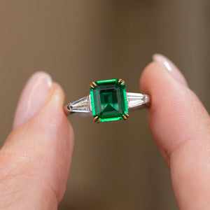 Handheld close-up of the emerald-cut centre stone framed by two bright trapezoid diamonds in white gold.