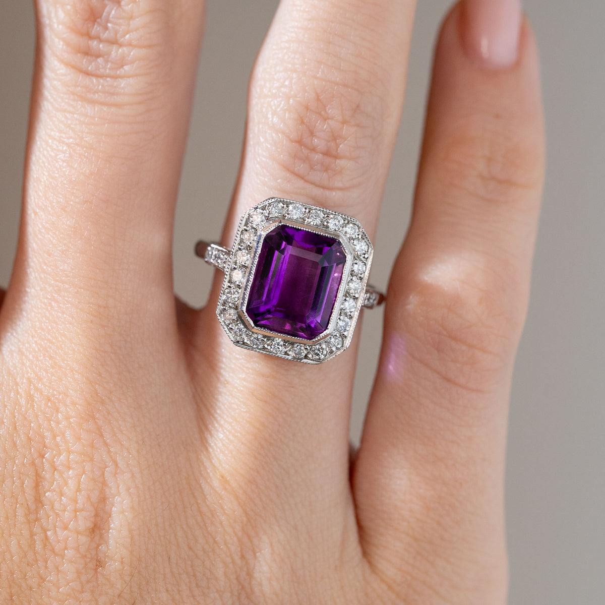 Close-up of an antique Art Deco amethyst and diamond cluster ring worn on a finger, featuring a vivid purple emerald-cut amethyst surrounded by round diamonds and delicate milgrain detailing.