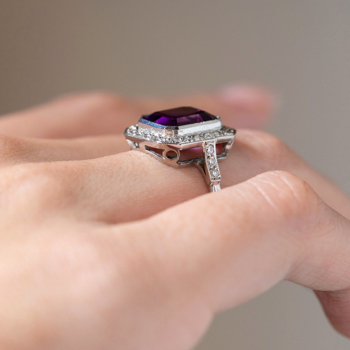 Side angle of an antique Art Deco amethyst and diamond cluster ring worn on a finger, revealing its raised rectangular setting, diamond halo and decorative white gold gallery.