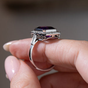 Side profile of an antique Art Deco amethyst and diamond cluster ring held between fingers, showing the elevated white gold setting, decorative open gallery and diamond-set shoulders.