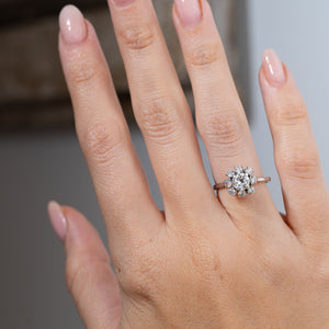 Vintage 1950s diamond cluster engagement ring worn on a hand, displaying its sparkling central diamond surrounded by eight smaller diamonds in a flower-shaped white gold setting.