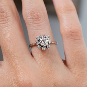 Close-up of a vintage 1950s diamond cluster engagement ring worn on a finger, highlighting the central round diamond and surrounding diamonds in an elegant white gold floral setting.