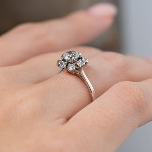 Side angle of a vintage 1950s diamond cluster engagement ring worn on a finger, revealing its raised diamond setting, detailed claw arrangement and slender white gold band.