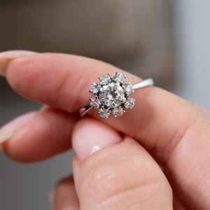 Detailed close-up of a vintage 1950s diamond cluster engagement ring held between fingers, highlighting the central round diamond surrounded by eight smaller diamonds in a symmetrical floral design.