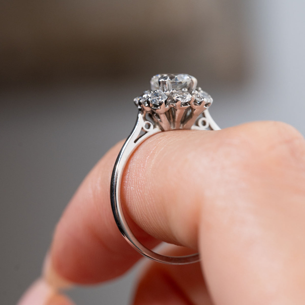 Side profile of a vintage 1950s diamond cluster engagement ring held between fingers, showcasing the elevated diamond cluster, decorative openwork gallery and polished white gold band.