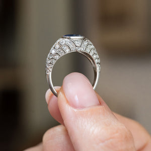 Reverse profile of an antique sapphire bombé ring held between the fingers, showing its curved platinum gallery and diamond-set shoulders.