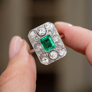 Colombian emerald and diamond ring held between the fingers, revealing the ornate geometric cluster and milgrain borders.
