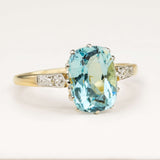 Antique Edwardian cushion-cut aquamarine and diamond engagement ring displayed on an off-white background, highlighting its craftsmanship and gemstone colour.