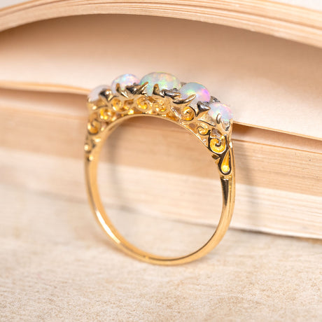 A yellow gold ring with opal stones set with diamonds in yellow gold, showing engraved scrolls on the setting on a book background. 