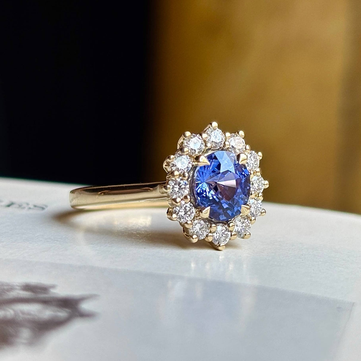 Blue sapphire and diamond cluster engagement ring, angled close-up on light surface showing fine details.