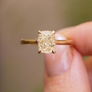 Antique-inspired, cushion-cut diamond solitaire engagement ring in yellow gold held in between the fingers against a blurry background.