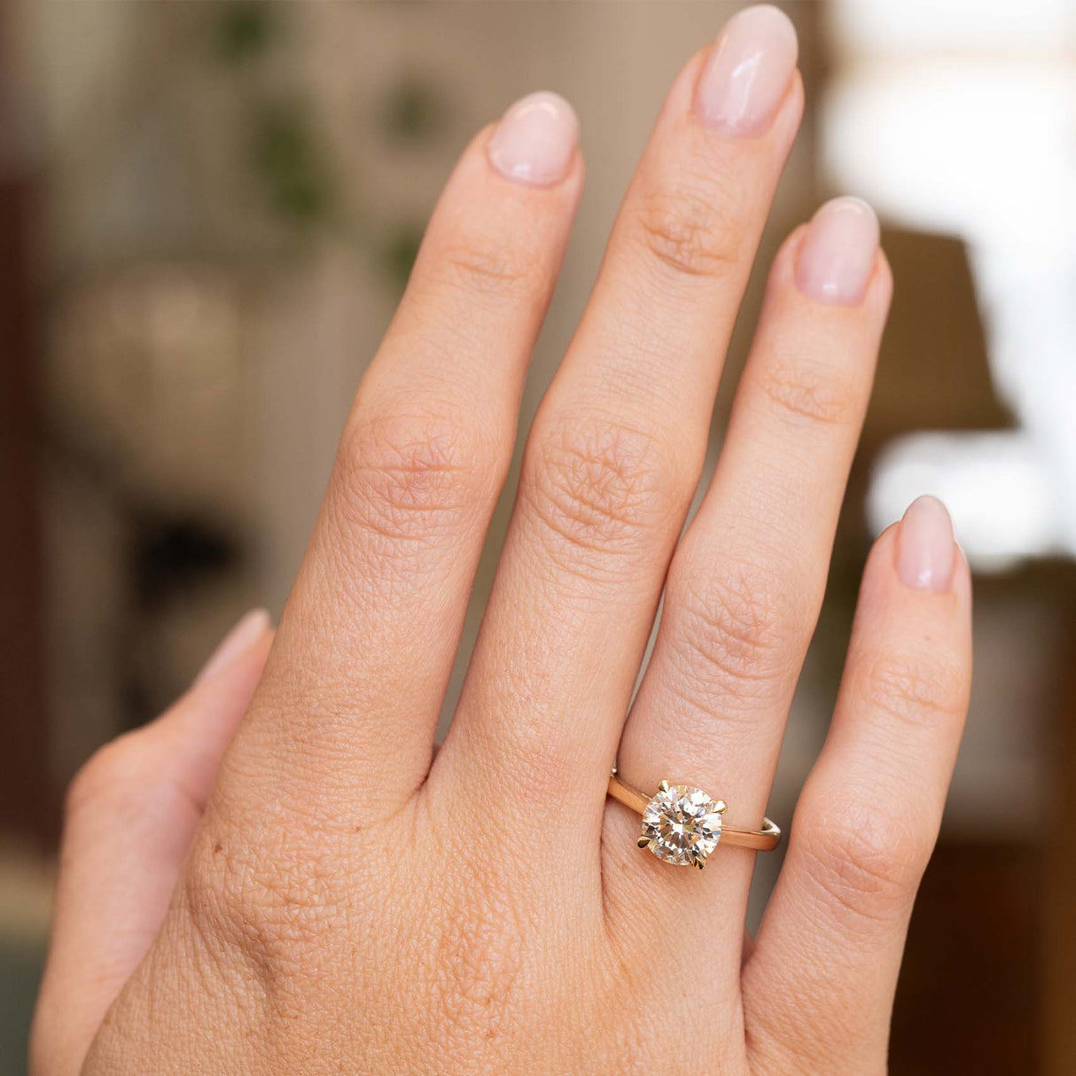 A close-up of a hand wearing a yellow gold ring with a round brilliant-cut diamond. The background is a softly blurred indoor setting.