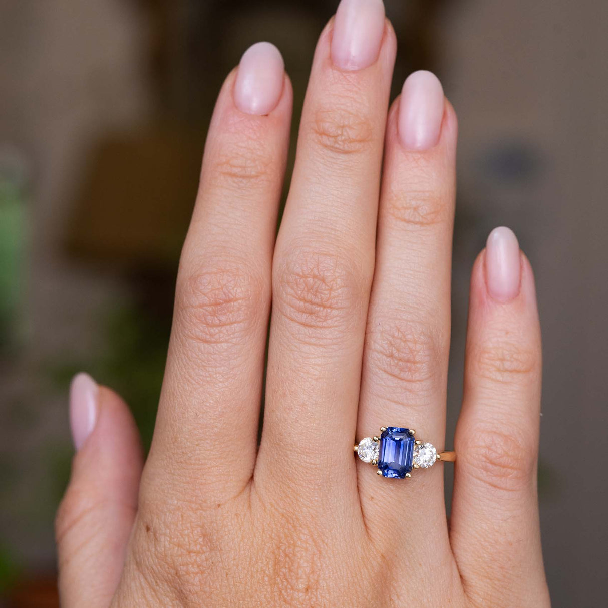 A close-up of a hand wearing an Antique-inspired, blue sapphire and diamond three-stone engagement ring with a yellow gold band set against a softly blurred background.