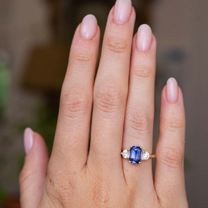 A close-up of a hand wearing an Antique-inspired, blue sapphire and diamond three-stone engagement ring with a yellow gold band set against a softly blurred background.