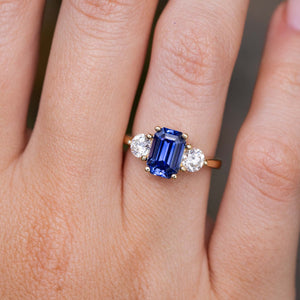 A close-up of a yellow gold three-stone engagement ring with a rectangular blue sapphire centre stone and two round clear diamonds on a finger. Inspired by antique designs.