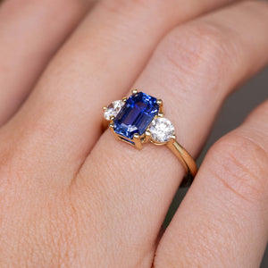 A close-up of a three-stone vintage inspired engagement ring.  Showcasing the central blue sapphire, two round diamond accent stones, and yellow gold band worn on a persons finger. 