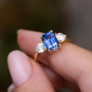 A close-up of a hand holding a vintage inspired three-stone engagement ring with a yellow gold band, central blue Sri Lankan sapphire and two flanking round-brilliant diamonds, set against a blurred natural background.