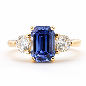 Front profile of a yellow gold three-stone engagement ring featuring an elongated rectangular blue sapphire centre stone and two round brilliant-cut diamonds on either side, set against a clean white background.