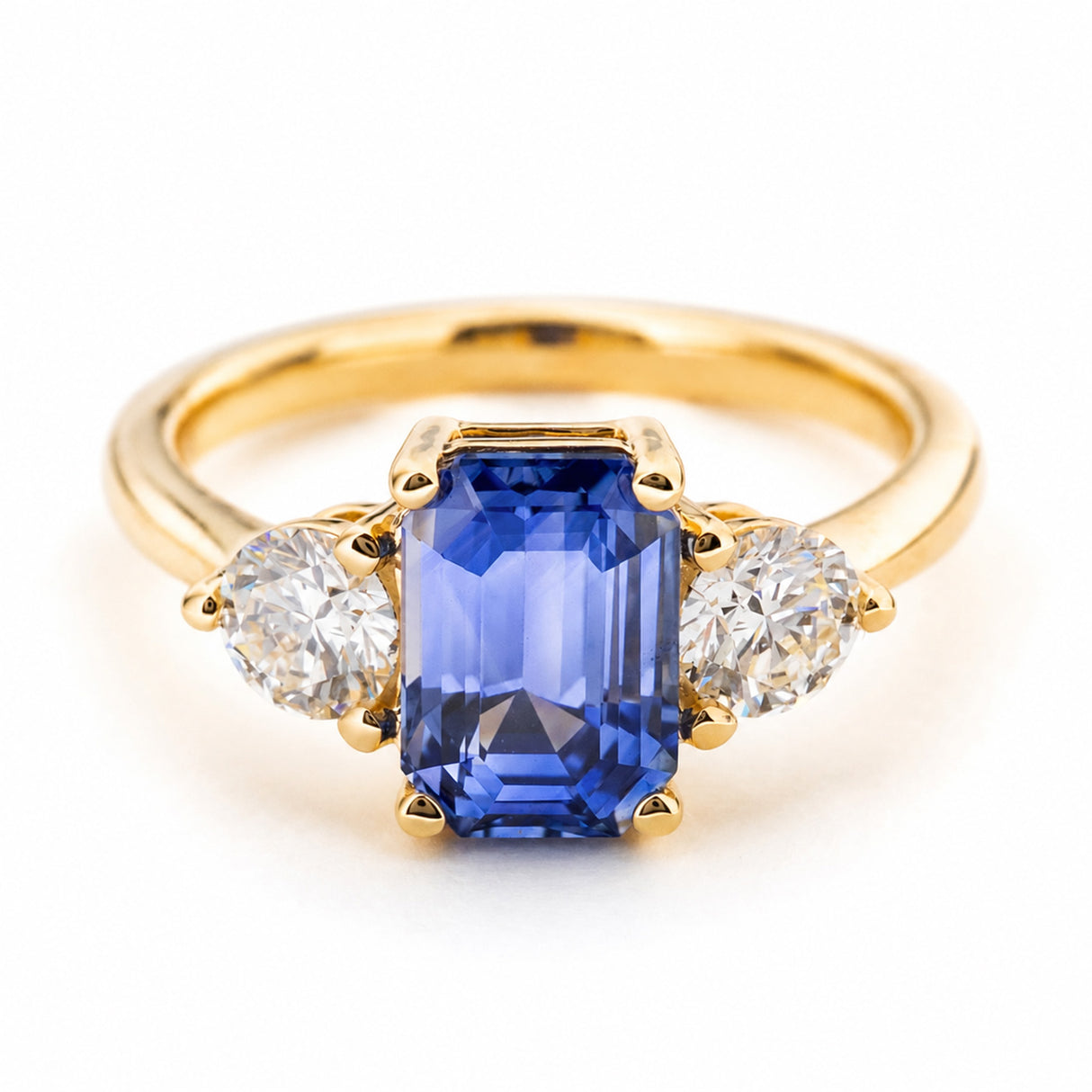A Vintage inspired three-stone engagement ring featuring a large rectangular Sri Lankan blue sapphire and two round clear diamonds on either side, with a yellow gold band set against a plain white background.