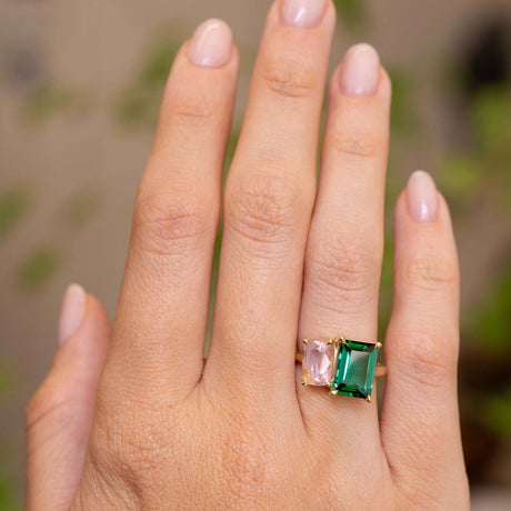 A close-up of a hand wearing a gold Vintage-inspired, Green tourmaline and pink sapphire Two stone engagement ring, set against a softly blurred outdoor background.
