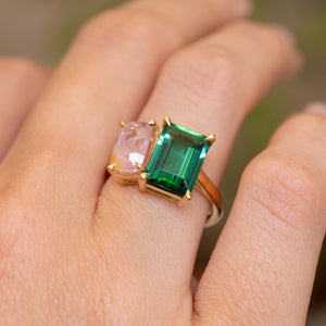 A close-up of a yellow gold Vintage style Green tourmaline and pink sapphire Toi et Moi engagement ring worn on a person's finger.