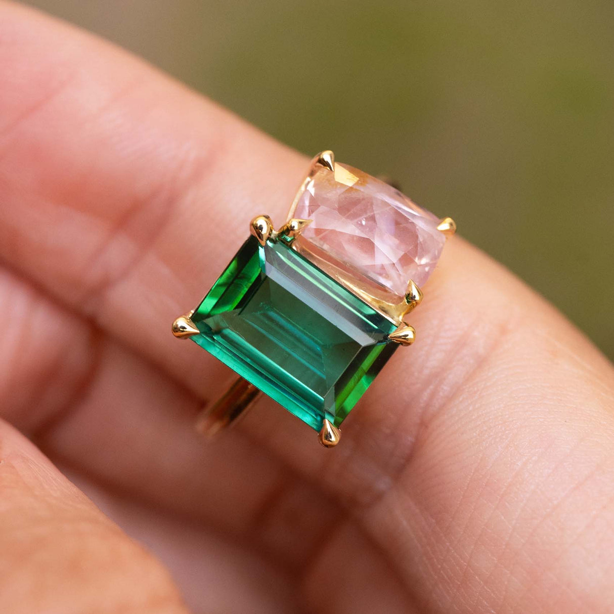 A gold ring featuring a large emerald-cut green tourmaline and a smaller pink sapphire shown worn on a person's finger against a soft blurred green background.