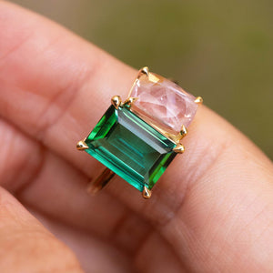 A gold ring featuring a large emerald-cut green tourmaline and a smaller pink sapphire shown worn on a person's finger against a soft blurred green background.