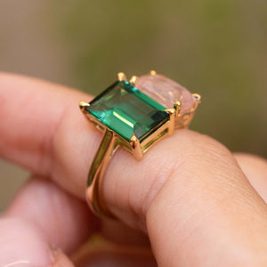 A yellow gold ring with a large emerald-cut green tourmaline and a pink sapphire  worn on a person's finger against a blurred green background