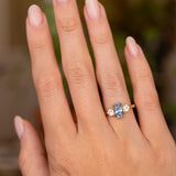 Cushion-cut blue sapphire engagement ring flanked by two round brilliant-cut diamonds, worn on the hand.