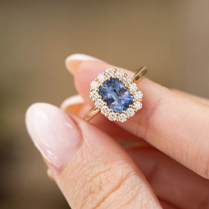 Antique-inspired blue sapphire engagement ring held between the fingers, displaying the cushion-cut centre stone and round brilliant diamond surround.