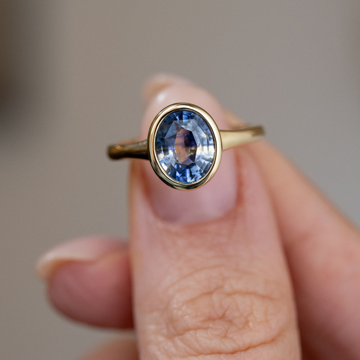Close fingertip view highlighting the sapphire’s shifting blue tones and polished yellow gold rub-over setting.