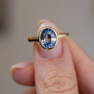 Close fingertip view highlighting the sapphire’s shifting blue tones and polished yellow gold rub-over setting.
