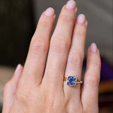 An image of an antique-inspired blue cushion cut sapphire bezel set in a yellow gold solitaire ring worn on the finger and shown at a distance against a blurred background.