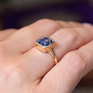 A close-up picture of an Antique-inspired blue sapphire bezel-set in a yellow gold solitaire ring worn on a finger and shown at an angle to display the side profile of the ring.