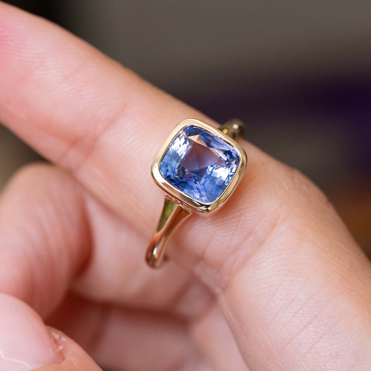 A close-up image of an antique-inspired cushion-cut blue sapphire solitaire ring in yellow gold positioned on a finger against a blurred background.