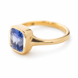 An antique-inspired blue sapphire bezel set solitaire ring in yellow gold and turned at an angle to show the shoulders of the ring and depth of the bezel, seen against a white background.