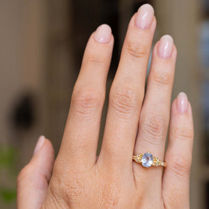 A close-up of a hand wearing a yellow gold vintage-inspired engagement ring with a central oval blue sapphire and small side diamonds, set against a softly blurred neutral background.