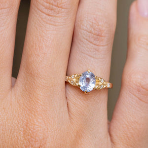 A close-up of a hand wearing a yellow gold engagement ring that is inspired by vintage designs.  Set  with an oval light blue sapphire and six small diamonds. 