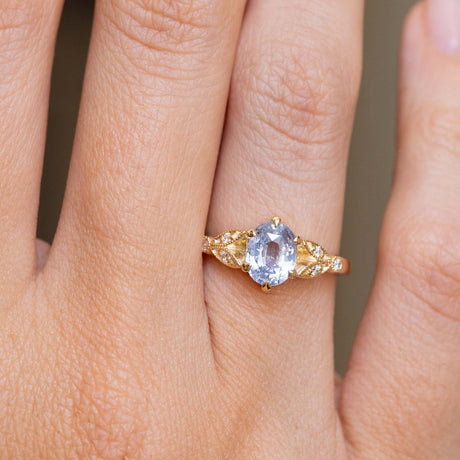 A close-up of a hand wearing a yellow gold engagement ring that is inspired by vintage designs.  Set  with an oval light blue sapphire and six small diamonds. 