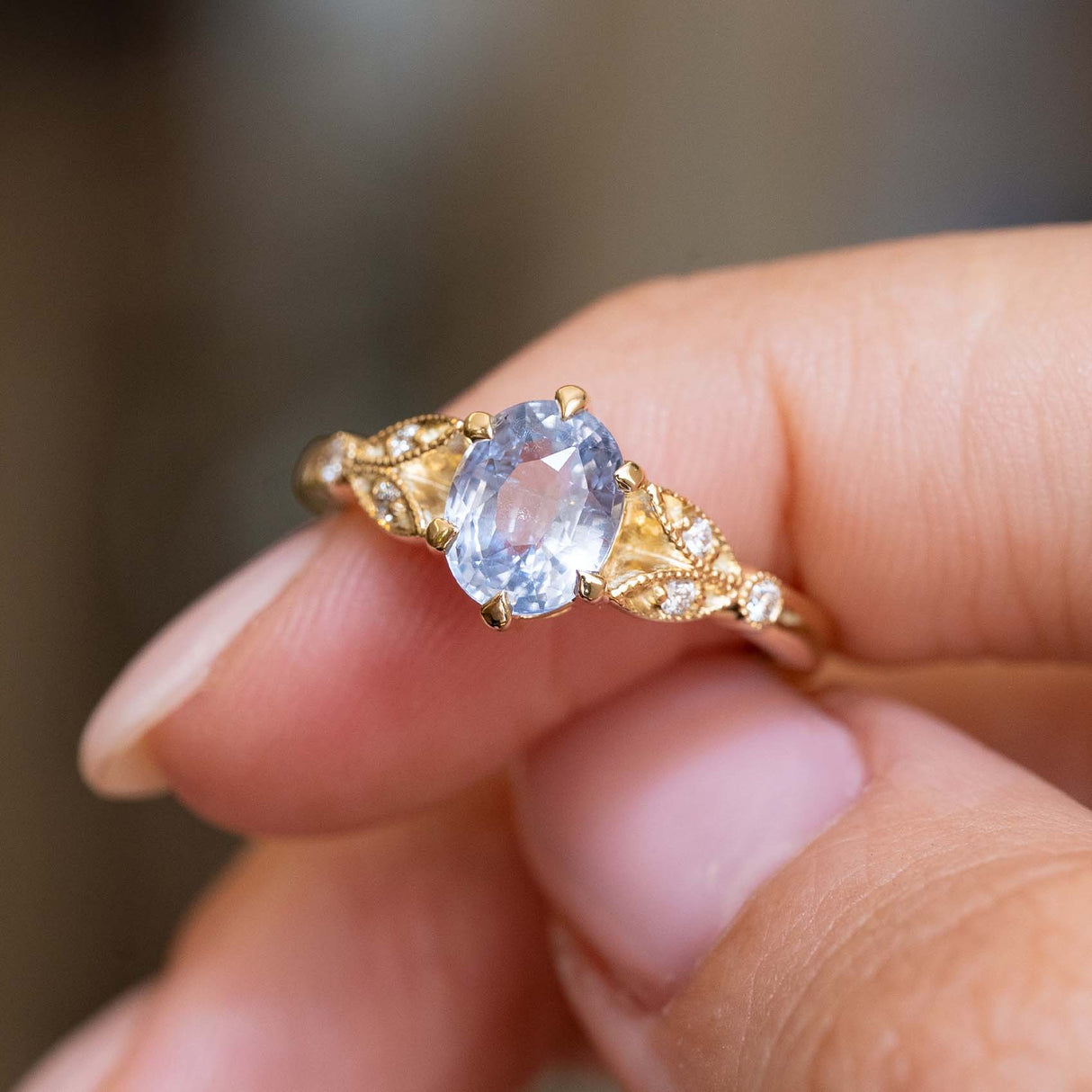 A close-up of a Vintage-inspired, yellow gold engagement ring with an oval light blue central sapphire and diamond accent stones, held between fingers against a softly blurred background.