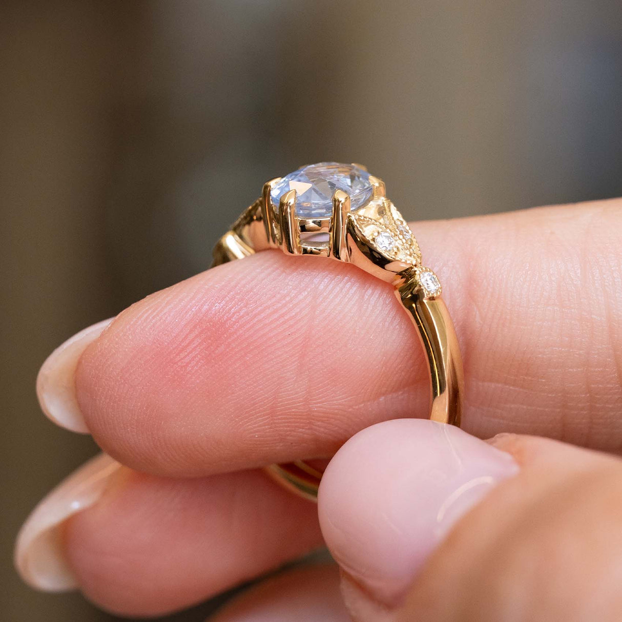 A close-up of a hand holding a yellow gold engagement ring with a large pale blue oval-cut sapphire and decorative side details. Vintage-inspired. 