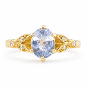 A Vintage-inspired,  yellow gold engagement ring featuring an oval-cut light blue sapphire at the centre, flanked by decorative gold filigree sections with small clear accent diamonds, set against a plain white background.