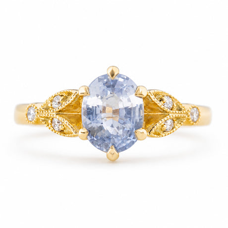 A Vintage-inspired,  yellow gold engagement ring featuring an oval-cut light blue sapphire at the centre, flanked by decorative gold filigree sections with small clear accent diamonds, set against a plain white background.