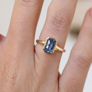 Close finger detail highlighting the cool blue sapphire and warm yellow gold bezel in a clean geometric design.