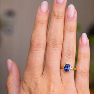 A close-up of a hand wearing an Antique-inspired, Blue sapphire solitaire engagement ring in yellow gold, set against a softly blurred neutral background.