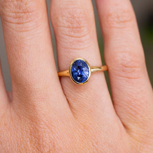 A yellow gold Antique-inspired solitaire engagement ring with an oval-cut deep blue sapphire and yellow gold band, shown worn on a person's finger.