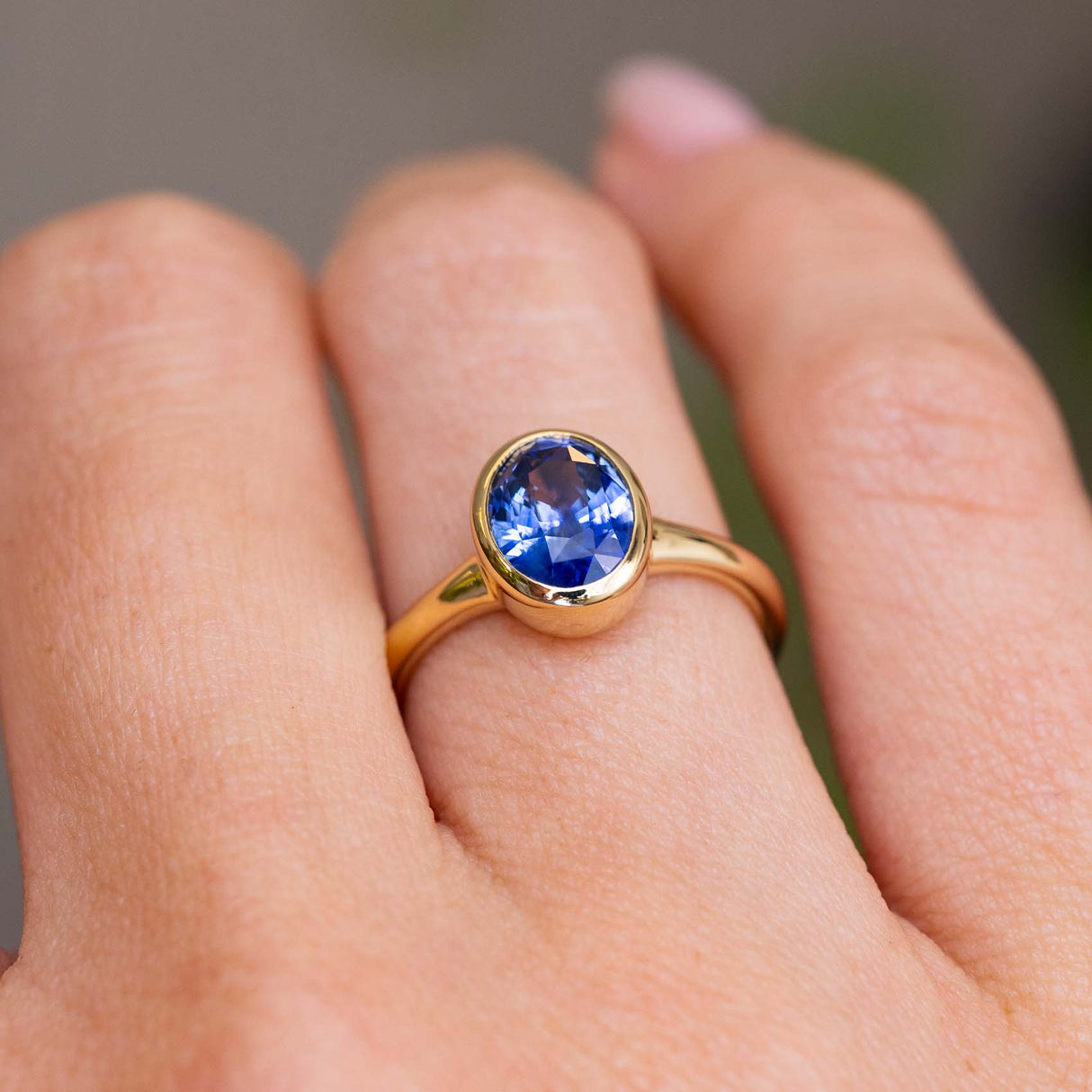 A solitaire engagement ring with a blue violet oval sapphire in a yellow gold bezel setting with a gold band.  Inspired by antique designs, worn on a finger and shown in a close-up lifestyle shot.
