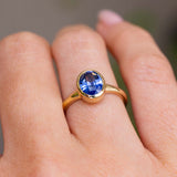 A solitaire engagement ring with a blue violet oval sapphire in a yellow gold bezel setting with a gold band.  Inspired by antique designs, worn on a finger and shown in a close-up lifestyle shot.